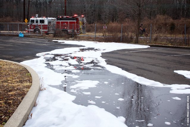 Parking lot with extinguishing foam and water on the ground, two fire trucks with lights on in the background near a fence and trees.