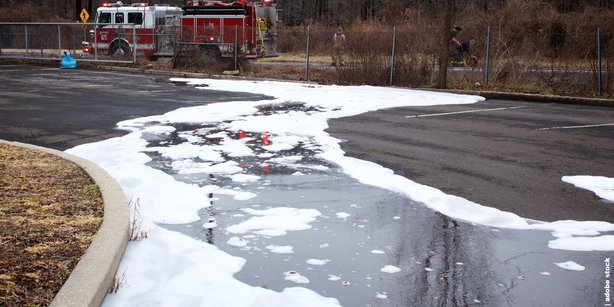 Parking lot with extinguishing foam and water on the ground, two fire trucks with lights on in the background near a fence and trees.