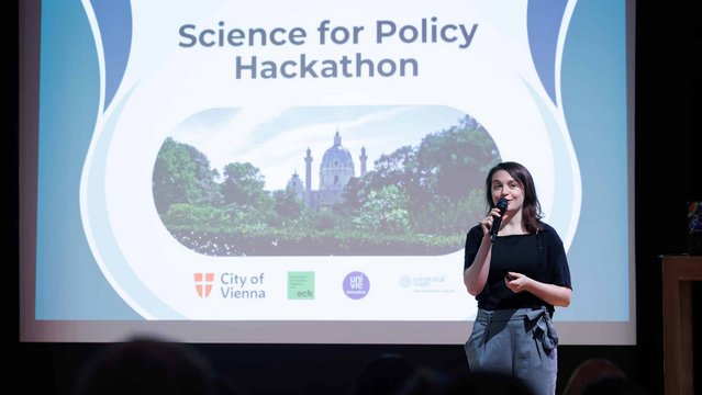 The moderator for the Hackathon Pitch day standing in fron of a powerpoint presentation on the stage, welcoming the audience