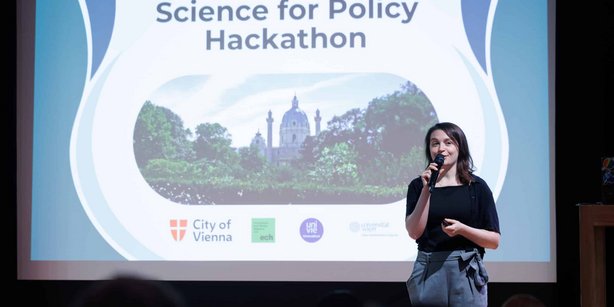 The moderator for the Hackathon Pitch day standing in fron of a powerpoint presentation on the stage, welcoming the audience