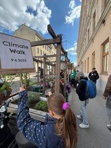 Eine Gruppe von Menschen geht auf einem Gehweg neben einem Gebäude entlang. Eine Person im Vordergrund hält ein Schild mit der Aufschrift „Climate Walk 9.4.2025“. Der Himmel ist teilweise bewölkt, und im Hintergrund sind Gebäude und Pflanzen zu sehen.