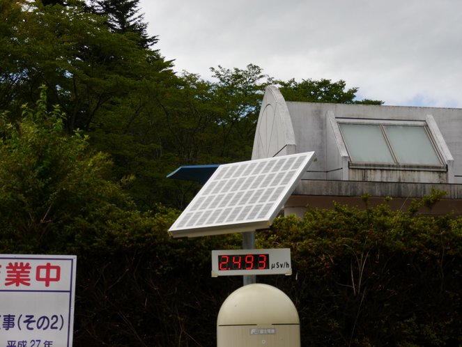 Solar-powered radiation measuring device in Namie displaying 2.493 µSv/h, with trees and a building in the background.