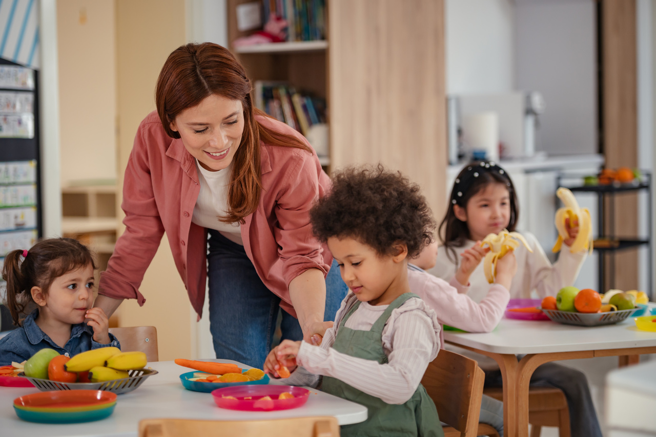 A Kindergarden group with three children eating and an adult (the teacher) talking to them