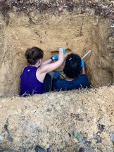 Two reserachres in a pit in the ground, getting soil samples for the project