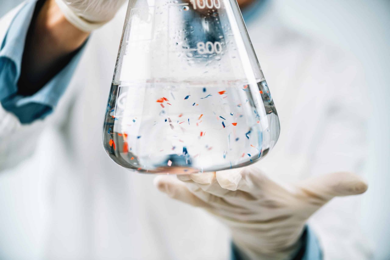 Gloved hands holding a 1000 ml flask with clear liquid and small colored particles, used for plastic pollution research.