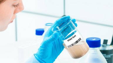Gloved hand holding a beaker labeled 'PFAS Test' with brown liquid inside; lab equipment visible in the background.