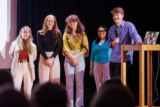 A team of five people presenting their pitch, with one member standing at the front, all of them smiling ot the audience
