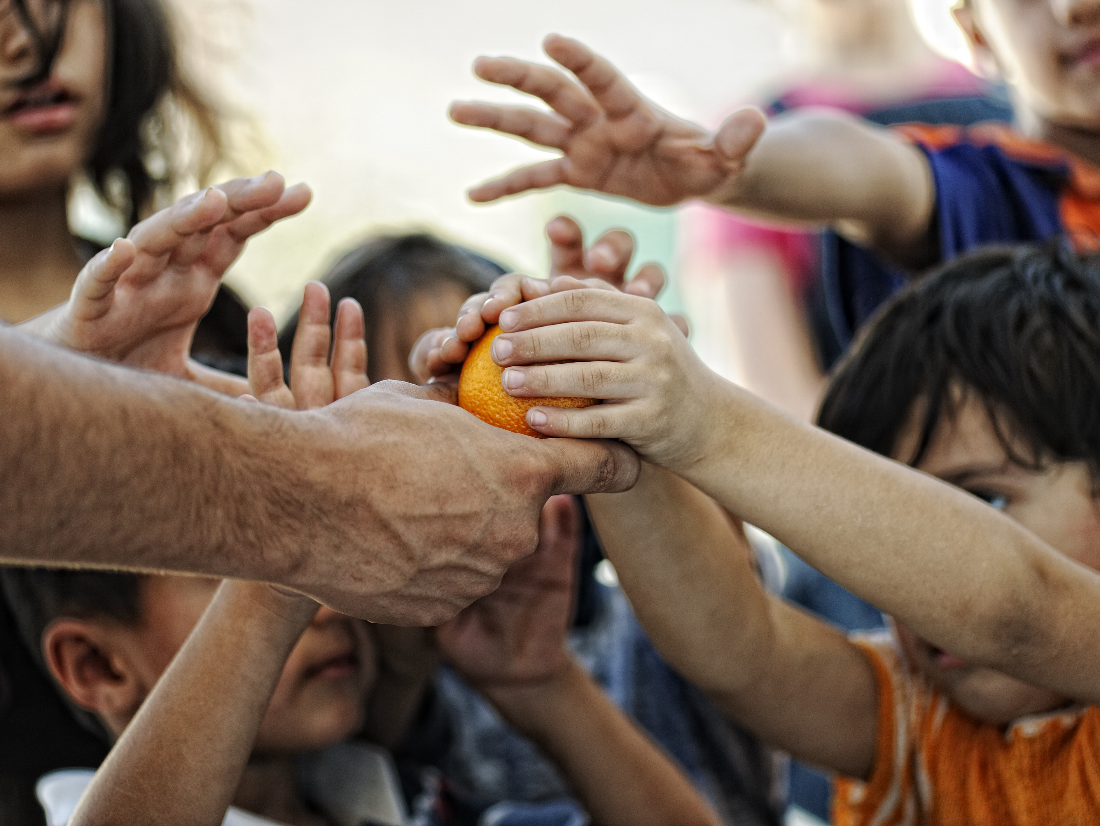 An adult hand handing an orange to a group of children reaching for it