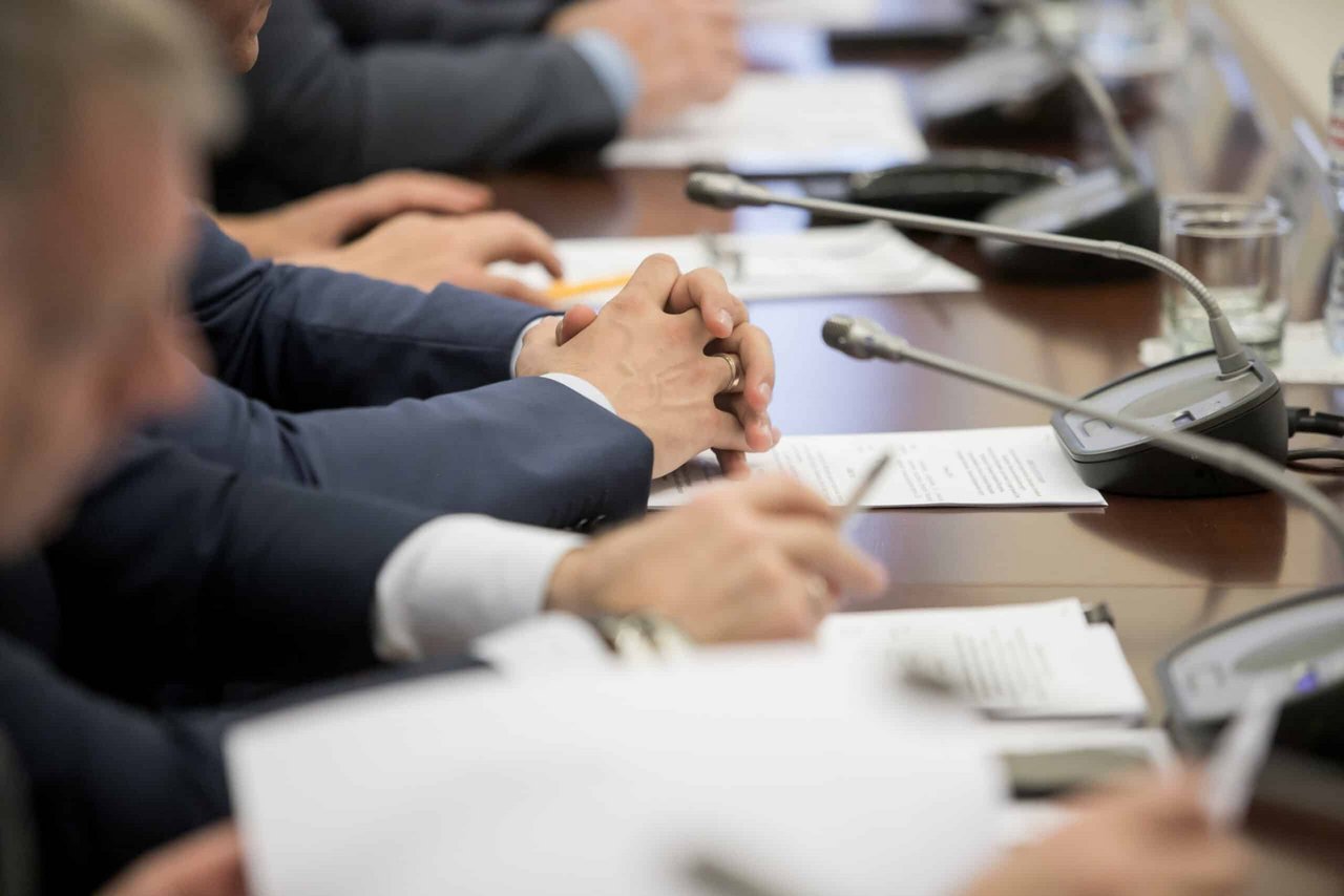 Group of people seated at a conference table with microphones, pens, and documents in front of them.