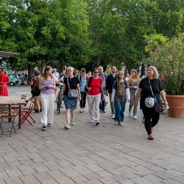 Eine Gruppe von Menschen geht auf einem gepflasterten Weg entlang eines Gewässers spazieren. Die Szene spielt sich in einem Außenbereich mit üppigen grünen Bäumen und Pflanzen ab. Einige Menschen gehen paarweise, während andere allein unterwegs sind. Auf der linken Seite befinden sich Bänke und Tische, und auf der rechten Seite nahe dem Wasser stehen große Topfpflanzen.