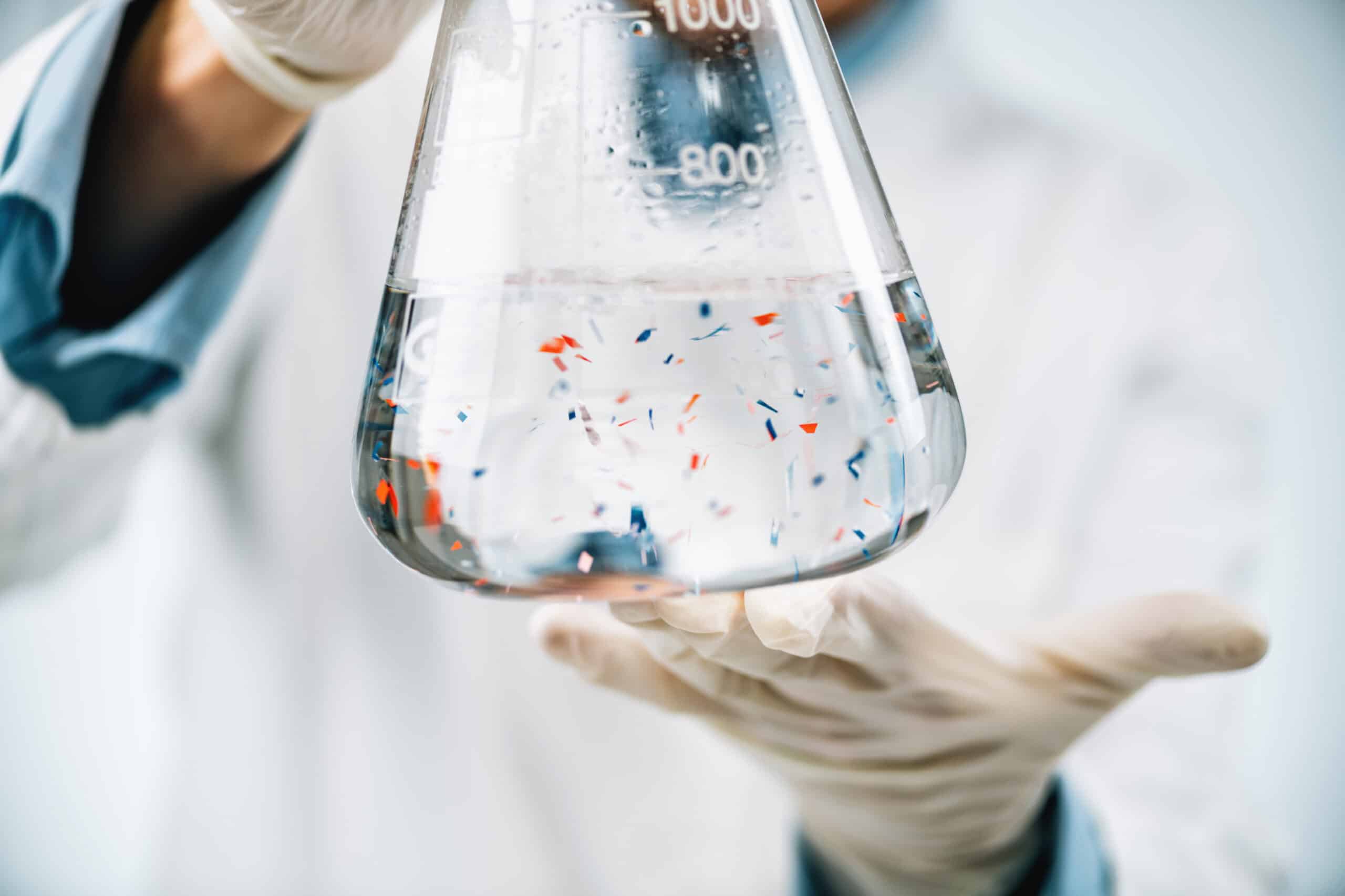Gloved hands holding a 1000 ml flask with clear liquid and small colored particles, used for plastic pollution research.
