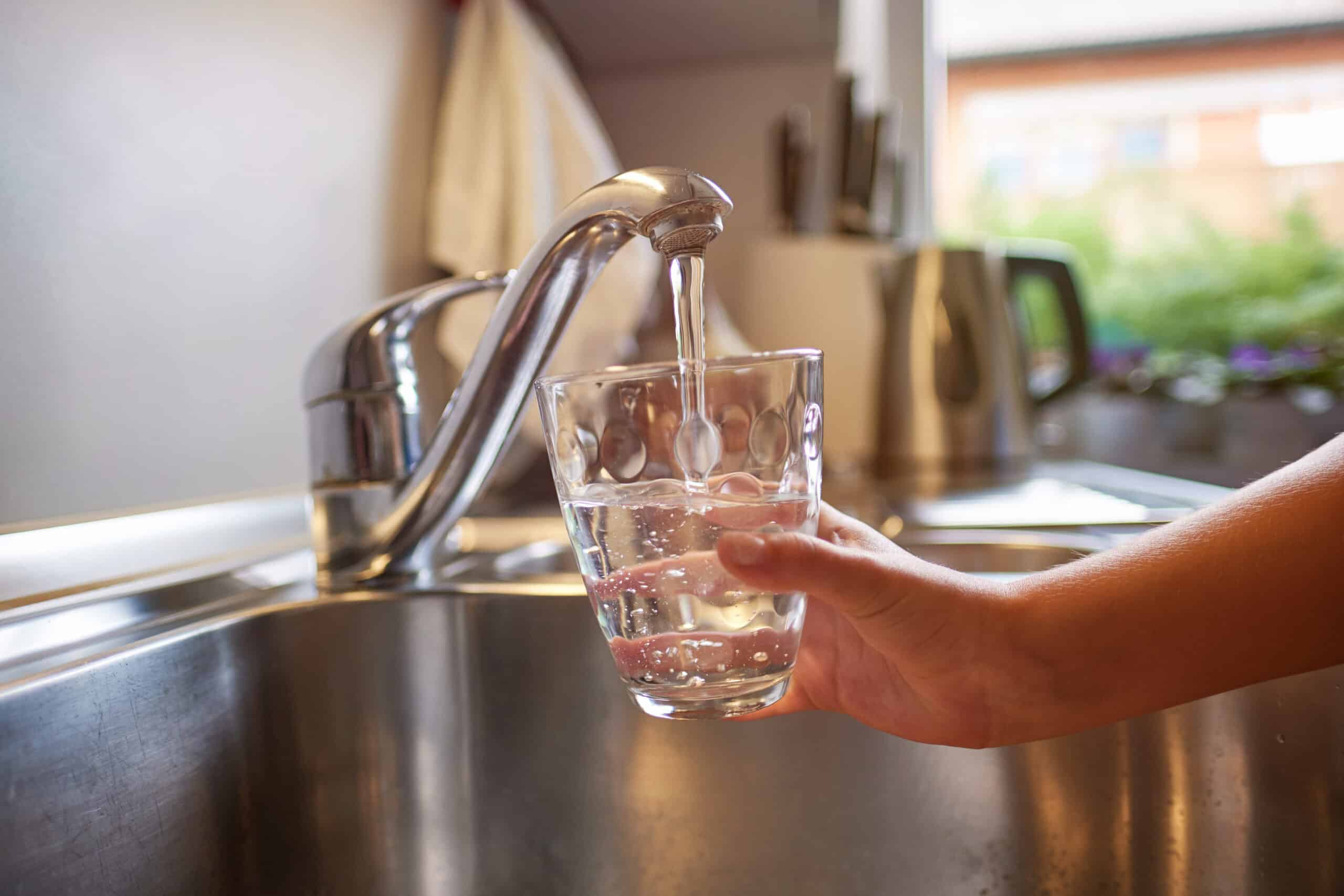 Hand holding a glass under a kitchen faucet with water flowing into it, in a kitchen setting with sink, kettle, and plants.