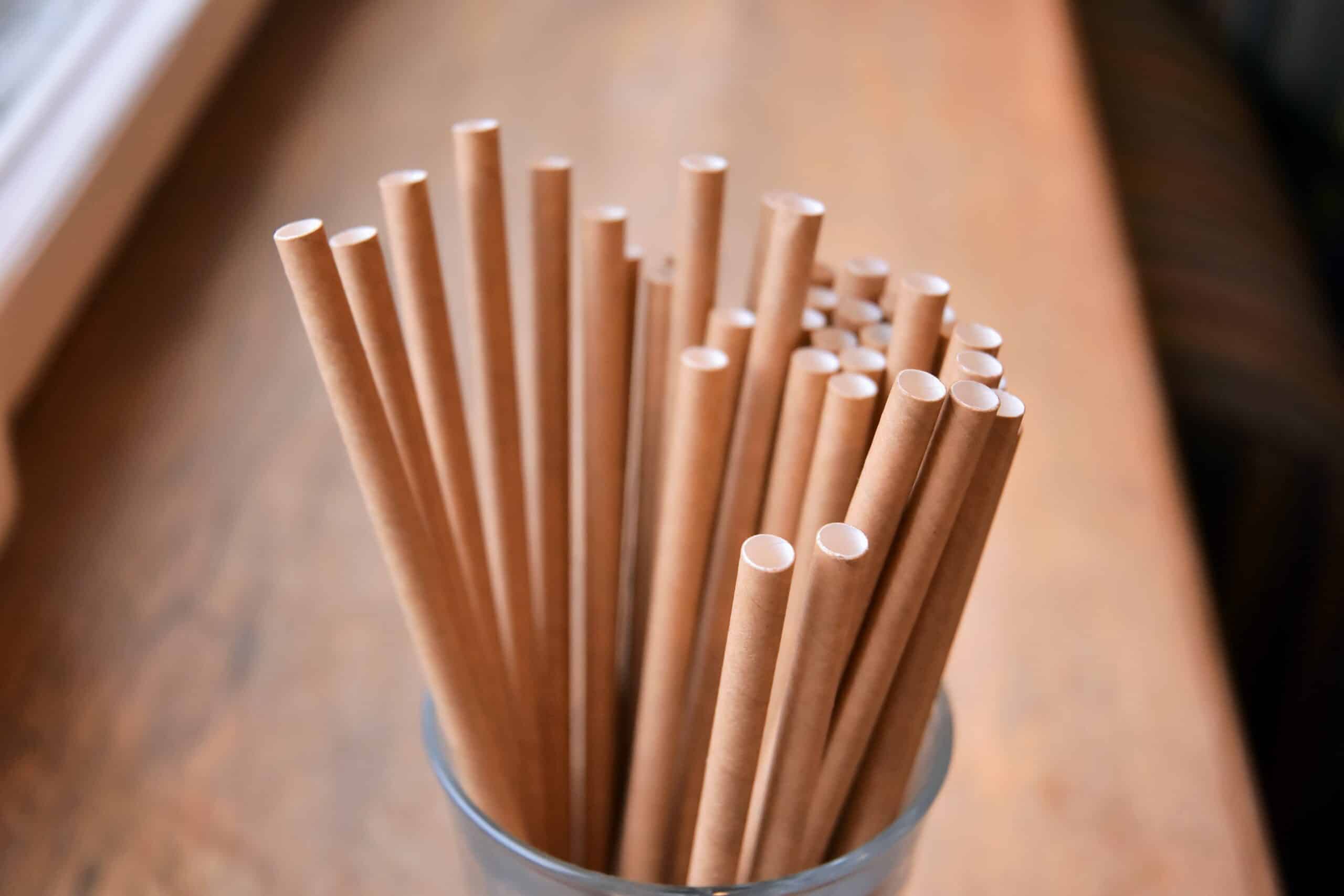 Close-up of brown paper straws standing upright in a glass container on a wooden surface.